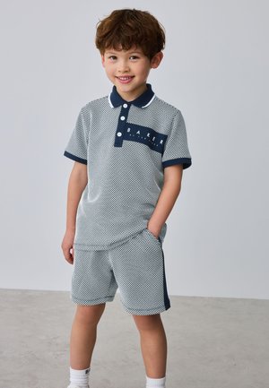 Junger Junge lächelt, trägt ein kurzärmliges gemustertes Poloshirt und passende Shorts mit Seitenstreifen, Hände in den Taschen, steht vor einem einfarbigen Hintergrund.