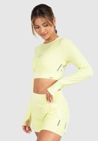 Helles gelbes Sportoutfit bestehend aus einem langärmeligen Cropped-Top und hoch taillierten Shorts, mit reflektierenden Akzenten und dehnbarem, glattem material.
