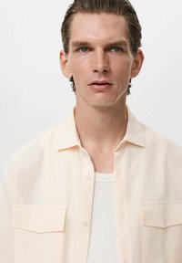 Lys beige kortærmet skjorte med spredt krave, to brystlommer og knaplukning foran. Bæres over en hvid ribbet tanktop. Glat tekstur.