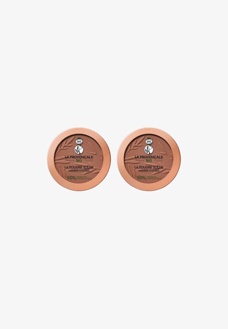 Deux compacts ronds de bronzer avec une poudre brun clair. Chacun a un couvercle de couleur bronze avec des détails de logo verts et noirs.