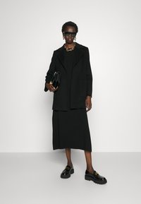 Sort skræddersyet blazer med spids krave, parret med en lang sort kjole. Accessories inkluderer en kraftig guldkæde-halskæde og solbriller. Sorte loafers.