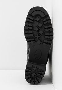 Semelle de chaussure en caoutchouc noir avec des rainures profondes, présentant un motif en croix et un détail de talon arrondi. Côté marqué de la taille "37" et du logo de la marque.