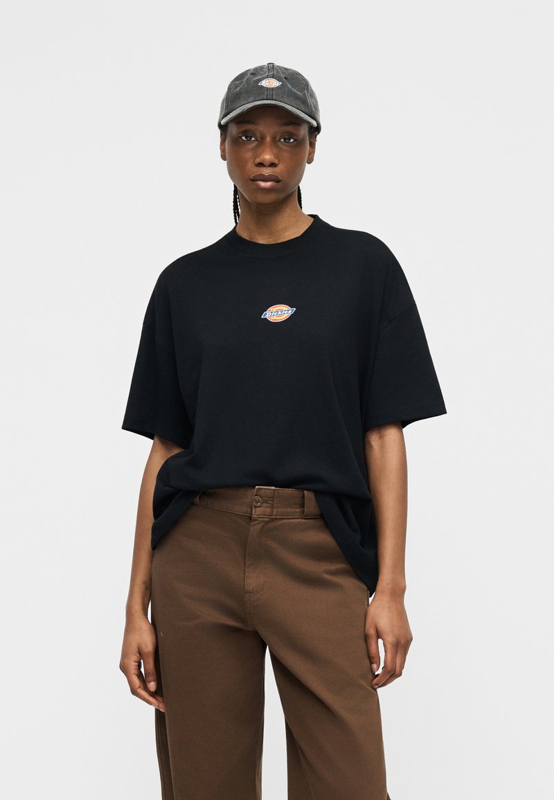 Donna che indossa una t-shirt nera oversize e pantaloni marroni, abbinati a un berretto grigio scuro, entrambi con il logo Dickies.