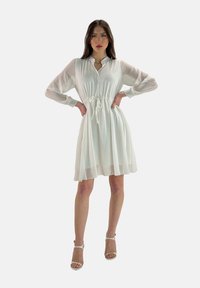 Vestido blanco hasta las rodillas con cierre de botones y cintura con cordón. Presenta mangas largas y transparentes y una tela ligera y en capas.