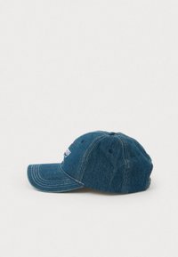 Niebieska denimsowa czapka baseballowa z widocznymi szwami, z czerwonym i białym haftowanym napisem częściowo widocznym z przodu, przedstawiona z boku na białym tle.