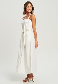 White cotton maxi dress with spaghetti straps, button front, and a tied waist. Includes side pockets and a flowing, textured fabric.