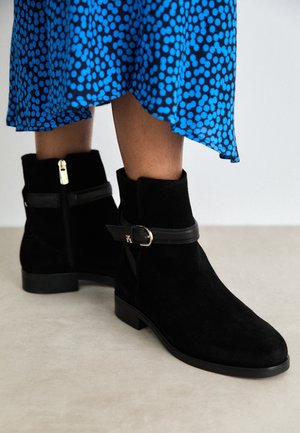 Jambes portant des bottines en suède noir avec fermetures éclair latérales et sangles à boucles, assorties à une robe bleue à pois noirs.