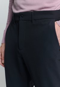 Marineblauwe broek met een gladde textuur, voorzien van een knoopsluiting, riemlussen en contrasterende stiksels. Close-up uitzicht met een lichte trui.