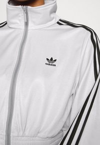 White zip-up jacket with high collar, black stripes along sleeves, elastic waistband, featuring a small black Adidas logo on chest.