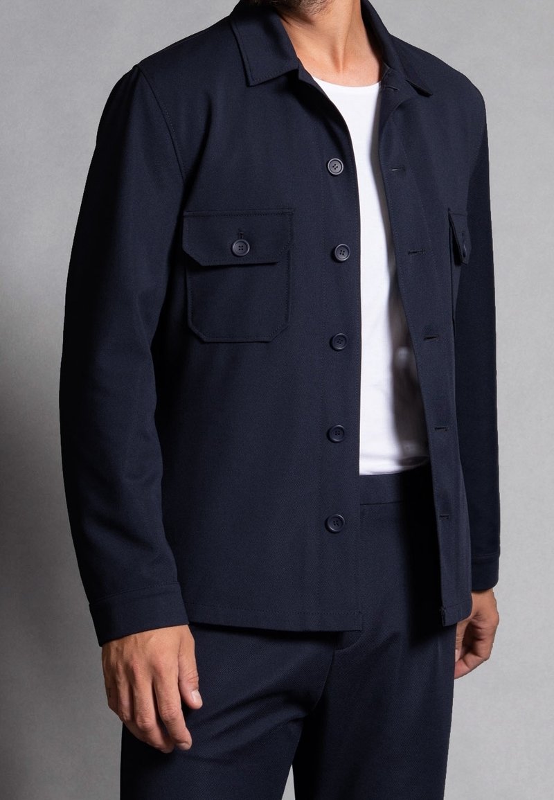 Navy blue jacket made from textured fabric, featuring a button-up front, two front pockets, and a pointed collar. Accompanied by a white shirt.