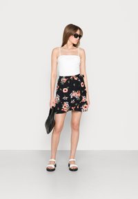 Saia mini preta com flores em rosa e branco, combinada com uma blusa de alças spaghetti branca e óculos de sol quadrados pretos, segurando uma bolsa preta.