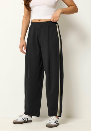 Kvinde iført sorte vidde bukser med beige sidelister, hvid cropped top og hvide sneakers mod en enkel lys baggrund.