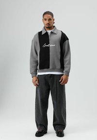 Grijs-zwart vest met ritssluiting en wijde mouwen, met een logo in het midden. Gecombineerd met losse zwarte jeans en zwarte schoenen.