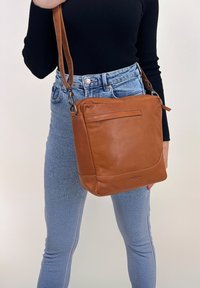 Bolso cruzado de cuero marrón con forma estructurada, un bolsillo frontal con solapa y herrajes plateados. Combinado con jeans y una blusa negra.