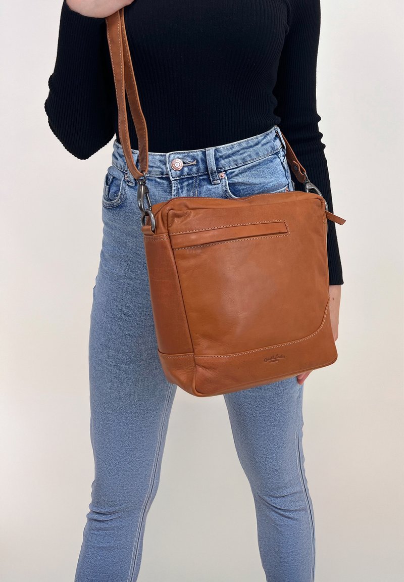 Bolso cruzado de cuero marrón con forma estructurada, un bolsillo frontal con solapa y herrajes plateados. Combinado con jeans y una blusa negra.