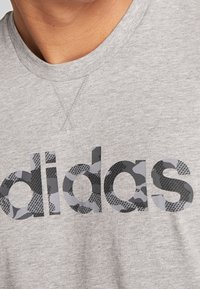 Grå T-shirt med kort ärm gjord av mjukt tyg, med en logotyp i svart och grå kamouflage tryckt över bröstet och rundad halsringning.