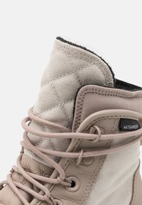 Detailní záběr na béžovou vodotěsnou botu s prošívaným jazykem, koženým lemem a odpovídajícími šněrovadly protaženými kovovými očky.