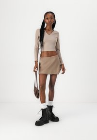 Béžový crop top s dlhým rukávom skombinovaný s priliehavou béžovou minisukňou. Modelka má na sebe čierne bojové topánky a drží malú hnedú kabelku.