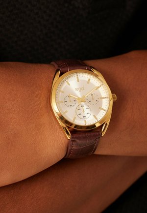 Gold wristwatch with white dial, three subdials, and brown leather strap worn on a person's wrist against a dark, textured background.