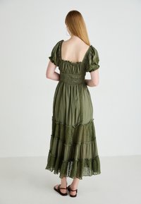 Olijfgroene maxi-jurk met pofmouwen, gerimpelde corsage en gelaagde kantdetails aan de zoom. Gepaard met bruine sandalen.