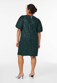 Abito verde smeraldo con paillettes, maniche a sbuffo, dettaglio con apertura a chiave sulla schiena e lunghezza fino al ginocchio. Indossato con sandali con tacco argento.