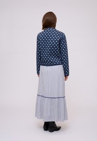 Donna con capelli castani lunghi fino alle spalle, che indossa una giacca blu a motivi, una lunga gonna a righe blu e bianche e stivali neri alla caviglia, di spalle.