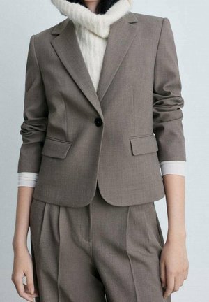 Femme portant un blazer brun ajusté avec des manches froncées, par-dessus un pull col roulé blanc, et un pantalon plissé assorti brun.