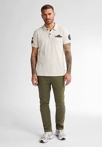 Tricou polo bej cu guler și mâneci scurte, având un buzunar maro pe piept și pantaloni verde măsliniu. Modelul stă pe un fundal neutru.