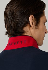 Mand med mørkt hår set fra siden, iført en navyblå skjorte med en rød krave, hvor ordet "HACKETT" står med mørke bogstaver.