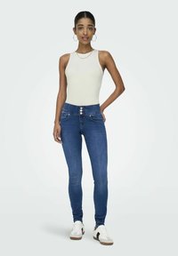 Slanke blå jeans med høj talje, sølvknaplukning og sidelommer; parret med en tætsiddende hvid tank top og casual sneakers.