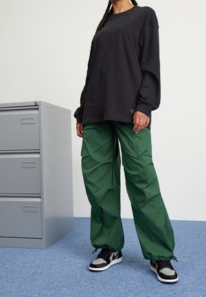 Chemise noire à manches longues associée à un pantalon cargo vert ample et des baskets noir et gris. L'arrière-plan présente un meuble de classement gris.