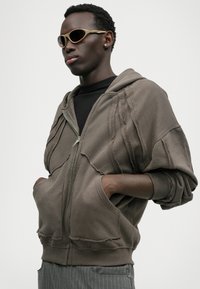 Brun oversized zip-up hoodie med slidte detaljer, forlommer og ribbede manchetter, parret med sort t-shirt og solbriller.