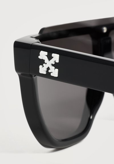 Lunettes de soleil noires à monture épaisse avec un logo blanc en forme de croix-flèche sur la branche et des verres teintés foncés.