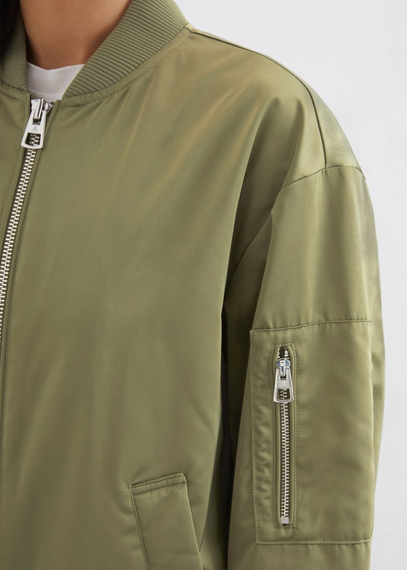 Olivegrüne Bomberjacke mit satinierter Oberfläche, ausgestattet mit ribbeltem Kragen, Frontreißverschluss und Taschenreißverschluss am Ärmel mit silbernem Zubehör.
