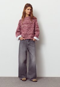 Pulover tricotat în nuanțe de roz, design dreptunghiular, decolteu rotund. Purtat cu blugi gri evazați și pantofi slip-on bej, modelul pozează pe un fundal simplu.