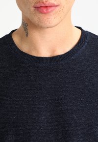 Suéter de punto azul marino con cuello redondo, tejido texturizado, ligero brillo y cuello acanalado; primer plano del área del escote.