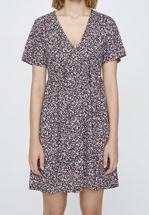 Femme portant une robe à manches courtes, col en V, longueur genou, avec un fond noir et un petit motif floral blanc et rose.