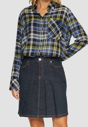 Žena nosící modrou, zelenou a černou kostkovanou košili na knoflíky zastrčenou do tmavé džínové plisované sukně s oranžovým prošitím.