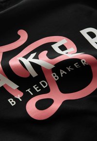 Musta kangas, jossa on rohkeat valkoiset ja vaaleanpunaiset kirjaimet. Tekstissä lukee ”TAKE” ja ”BY TED BAKER” näkyvillä, tyylitellyin kirjasimin. Sileä pinta.