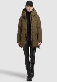 Olivgrüne gesteppte Puffjacke mit Kapuze, seitlichen Reißverschlüssen und schwarzen Akzenten, kombiniert mit glänzenden schwarzen Leggings und Kampfstiefeln.