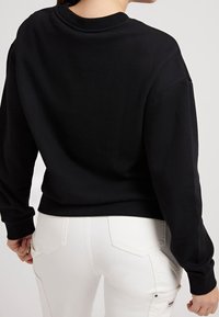Sudadera negra con cuello redondo acanalado, mangas largas y un corte relajado, combinada con pantalones blancos que tienen bolsillos traseros.
