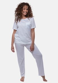 Nainen, jolla on kihara hiukset, hymyilee seisten paljain jaloin valkoisessa lyhythihaisessa paidassa, jossa on tasku, ja siihen sopivissa valkoisissa housuissa valkoista taustaa vasten.