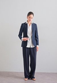 Granatowa marynarka dopasowana na białej koszuli, w połączeniu z ciemnymi, szerokimi spodniami. Gładki materiał, strukturalny krój i minimalistyczny styl.