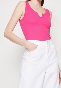 Dopasowany różowy prążkowany top na ramiączkach z falbaniastym dekoltem, zestawiony z białymi dżinsami o wysokim stanie, które mają boczne kieszenie i srebrne akcenty.