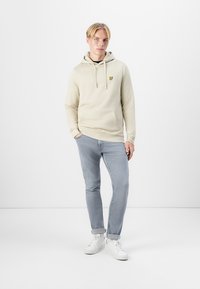 Hoodie beige clair avec une poche frontale, cordon de serrage réglable et petit logo jaune. Assorti avec un jean bleu clair et des sneakers blanches.