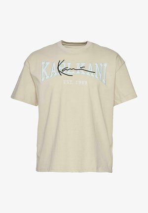 Beige puuvillane T-särk ümarate kaeltega, millel on rinnal sinine ja must tekst "KARL KANI" ning all aastat 1989.