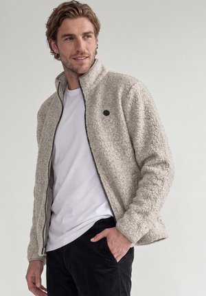 Homme portant une veste polaire beige clair sur une chemise blanche avec un pantalon noir, debout avec une main dans la poche, sur un fond uni.