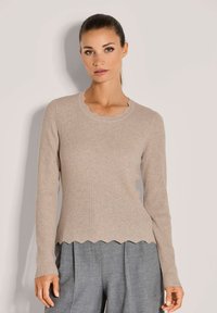 Frau in beiger, gerippter Sweater mit geschwungenen Kanten und grauen, plissierten Hose, die vor einem schlichten, hellen Hintergrund steht.