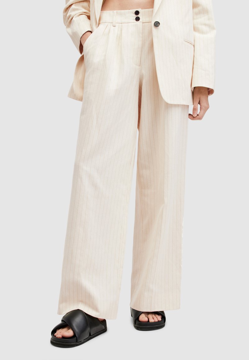 Pantaloni beige a righe con una vestibilità comoda, caratterizzati da un alto punto vita, due tasche frontali e un design sartoriale. Indossati con sandali neri a scivolo.