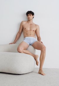 Helleblaue Herren-Boxershorts mit elastischem Bund, glattem Stoff und anliegendem Schnitt. Das Model sitzt auf einem weichen, geschwungenen Sofa.